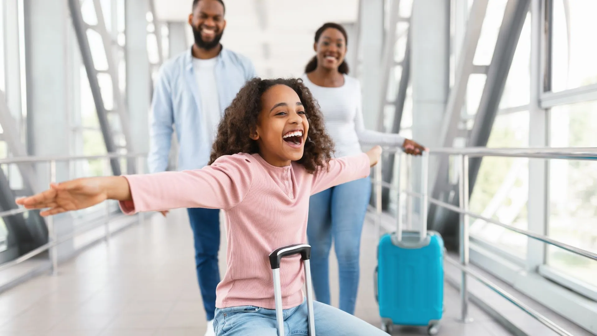 Uma menina negra de cabelos cacheados, vestindo uma blusa rosa, está sentada em sua mala e deslizando por um corredor de aeroporto com os braços abertos e uma expressão de pura alegria. Ao fundo, seus pais — um homem e uma mulher negros — caminham sorridentes, acompanhando a diversão da filha. O ambiente é moderno, com grandes janelas de vidro e estruturas metálicas, banhado por uma luz natural brilhante.