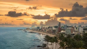 Uma fotografia panorâmica da orla de Salvador, na Bahia, sob o brilho dourado do pôr do sol. Em primeiro plano, no canto inferior direito, destacam-se as copas de vários coqueiros. A linha da costa segue em uma curva suave, com uma praia de areia clara interrompida por formações rochosas escuras que avançam sobre o mar azul. Ao longo da orla, há uma fileira de prédios residenciais e comerciais de altura variada. Ao fundo, em uma pequena península, vê-se a silhueta do Farol da Barra. O céu está preenchido por nuvens densas e expressivas, iluminadas por tons intensos de laranja e amarelo.