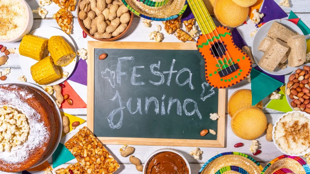 Uma mesa decorada de Festa Junina com fundo de madeira branca. Ao centro, há uma pequena lousa de giz verde com a inscrição "Festa Junina". Ao redor, estão dispostos pratos e tigelas com comidas típicas brasileiras: espigas de milho cozido, bolo de fubá com pipoca, pé de moleque, amendoim com e sem casca, paçoca, canjica salpicada com canela e doce de leite. A decoração inclui bandeirinhas coloridas, minichapéus de palha e um pequeno violão de brinquedo decorado em tons de laranja e verde.