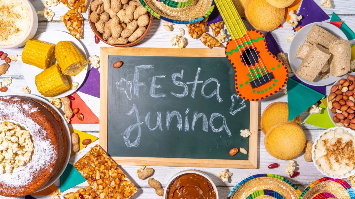 Uma mesa decorada de Festa Junina com fundo de madeira branca. Ao centro, há uma pequena lousa de giz verde com a inscrição "Festa Junina". Ao redor, estão dispostos pratos e tigelas com comidas típicas brasileiras: espigas de milho cozido, bolo de fubá com pipoca, pé de moleque, amendoim com e sem casca, paçoca, canjica salpicada com canela e doce de leite. A decoração inclui bandeirinhas coloridas, minichapéus de palha e um pequeno violão de brinquedo decorado em tons de laranja e verde.