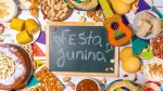 Uma mesa decorada de Festa Junina com fundo de madeira branca. Ao centro, há uma pequena lousa de giz verde com a inscrição "Festa Junina". Ao redor, estão dispostos pratos e tigelas com comidas típicas brasileiras: espigas de milho cozido, bolo de fubá com pipoca, pé de moleque, amendoim com e sem casca, paçoca, canjica salpicada com canela e doce de leite. A decoração inclui bandeirinhas coloridas, minichapéus de palha e um pequeno violão de brinquedo decorado em tons de laranja e verde.