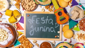 Uma mesa decorada de Festa Junina com fundo de madeira branca. Ao centro, há uma pequena lousa de giz verde com a inscrição "Festa Junina". Ao redor, estão dispostos pratos e tigelas com comidas típicas brasileiras: espigas de milho cozido, bolo de fubá com pipoca, pé de moleque, amendoim com e sem casca, paçoca, canjica salpicada com canela e doce de leite. A decoração inclui bandeirinhas coloridas, minichapéus de palha e um pequeno violão de brinquedo decorado em tons de laranja e verde.