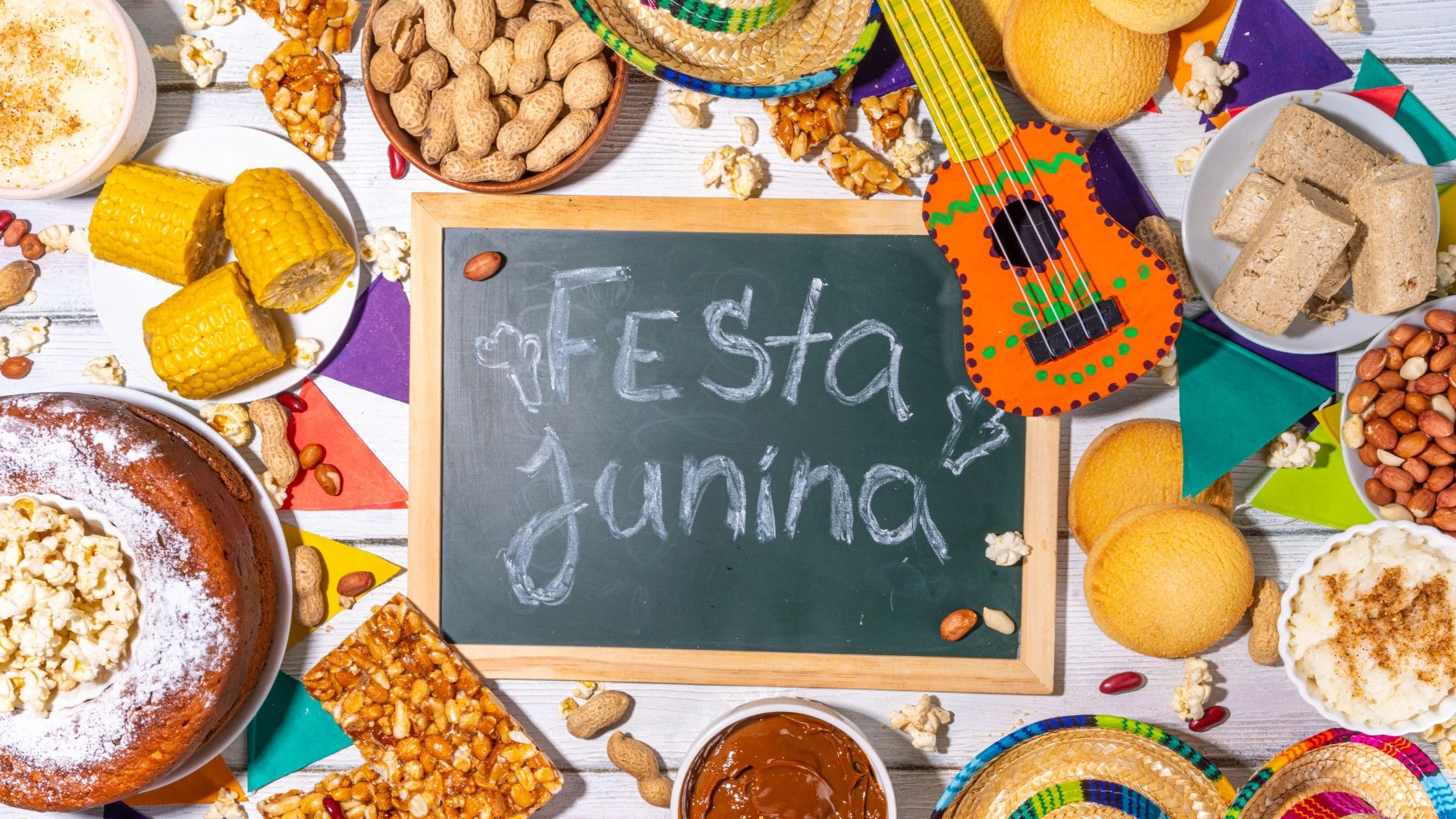 Uma mesa decorada de Festa Junina com fundo de madeira branca. Ao centro, há uma pequena lousa de giz verde com a inscrição "Festa Junina". Ao redor, estão dispostos pratos e tigelas com comidas típicas brasileiras: espigas de milho cozido, bolo de fubá com pipoca, pé de moleque, amendoim com e sem casca, paçoca, canjica salpicada com canela e doce de leite. A decoração inclui bandeirinhas coloridas, minichapéus de palha e um pequeno violão de brinquedo decorado em tons de laranja e verde.