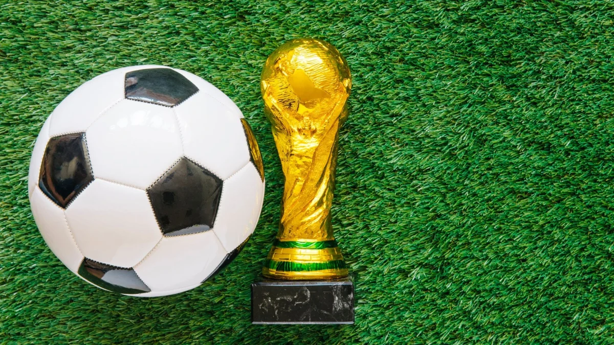 Vista aérea de uma bola de futebol clássica, em gomos brancos e pretos, posicionada ao lado esquerdo de uma réplica em miniatura do troféu da Copa do Mundo da FIFA. Ambos os objetos estão sobre um gramado sintético verde brilhante, que preenche todo o fundo da imagem.