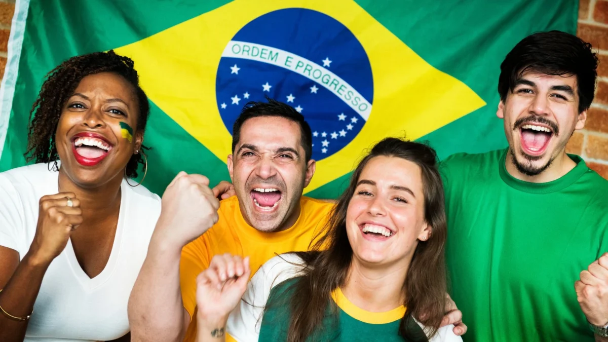 Quatro torcedores do Brasil felizes, sorrindo e vibrando em frente a uma bandeira nacional, que está pendurada em uma parede de tijolos. Uma mulher negra, um homem e uma mulher brancos e um homem de cabelos escuros estão juntos, com os braços levantados e as mãos em punho. A mulher negra e o homem de cabelos escuros estão rindo, com a boca aberta. A mulher negra tem tranças e usa um colar. O homem e a mulher brancos também sorriem. Eles vestem camisas verdes e amarelas, e a mulher negra tem a bandeira do Brasil pintada no rosto.