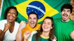 Quatro torcedores do Brasil felizes, sorrindo e vibrando em frente a uma bandeira nacional, que está pendurada em uma parede de tijolos. Uma mulher negra, um homem e uma mulher brancos e um homem de cabelos escuros estão juntos, com os braços levantados e as mãos em punho. A mulher negra e o homem de cabelos escuros estão rindo, com a boca aberta. A mulher negra tem tranças e usa um colar. O homem e a mulher brancos também sorriem. Eles vestem camisas verdes e amarelas, e a mulher negra tem a bandeira do Brasil pintada no rosto.