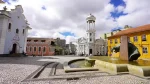 Uma fotografia em ângulo aberto de uma praça histórica e movimentada, pavimentada com calçamento de pedra irregular e detalhes em granito mais escuro, sob um céu azul brilhante e nuvens brancas fofas. No centro da praça, uma grande fonte de água em formato de U com uma escultura de uma gárgula jorrando água para o centro. Ao redor da praça, edifícios históricos de cores variadas: à esquerda, uma igreja de fachada rococó em branco e lilás claro com detalhes intrincados e um portal decorado com azulejos azuis e brancos; ao fundo, um edifício cor-de-rosa de telhado rústico; no centro-direita, um edifício neoclássico branco com uma torre campanário circular proeminente encimada por uma cúpula branca; e à direita, um edifício amarelo vibrante de dois andares com telhado de telhas vermelhas e janelas em arco. Pessoas estão caminhando, sentadas e andando de bicicleta pela praça, dando vida ao local. No horizonte, a paisagem urbana de edifícios modernos e montanhas distantes. O chão de pedras irregulares e a fonte de água em primeiro plano criam linhas diagonais fortes que levam o olhar para a profundidade da praça. A luz natural realça as texturas das pedras, dos edifícios e da água em movimento na fonte.