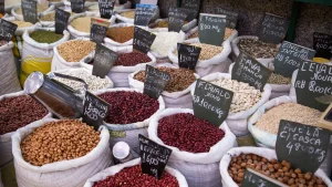 Uma fotografia em ângulo alto mostra dezenas de sacos de estopa abertos e preenchidos até a borda com grãos, nozes e leguminosas em uma banca de mercado. Em primeiro plano, os sacos contêm amendoins inteiros com casca, grãos de cacau vermelhos-escura, amendoins descascados e avelãs inteiras com casca. Sacos localizados mais para trás contêm vários tipos de feijão secos e brancos, grão-de-bico, sementes de girassol e flocos de aveia. Cada saco inclui uma pequena placa de identificação preta em português com a descrição do produto e o preço.