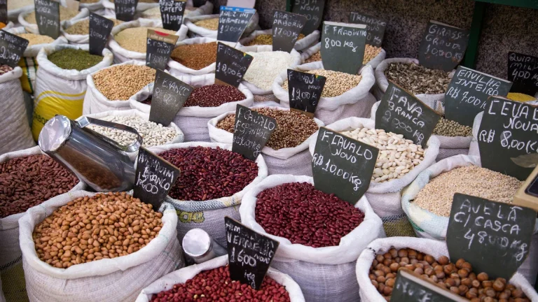 Uma fotografia em ângulo alto mostra dezenas de sacos de estopa abertos e preenchidos até a borda com grãos, nozes e leguminosas em uma banca de mercado. Em primeiro plano, os sacos contêm amendoins inteiros com casca, grãos de cacau vermelhos-escura, amendoins descascados e avelãs inteiras com casca. Sacos localizados mais para trás contêm vários tipos de feijão secos e brancos, grão-de-bico, sementes de girassol e flocos de aveia. Cada saco inclui uma pequena placa de identificação preta em português com a descrição do produto e o preço.