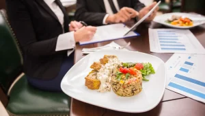 Uma imagem em close de um prato de comida em uma mesa de jantar durante uma reunião de negócios. O prato contém fatias de carne cobertas com um molho branco cremoso, acompanhadas por uma porção de arroz com vegetais e uma pequena salada lateral. Ao fundo, fora de foco, duas pessoas vestindo trajes formais (paletós) estão sentadas à mesa; uma delas escreve em um documento numa prancheta, enquanto a outra segura um tablet. Vários gráficos e planilhas estão espalhados sobre a mesa de madeira escura.