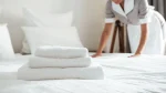 Uma pilha com três toalhas brancas e macias, dobradas de forma organizada, está em primeiro plano sobre uma cama com lençóis brancos impecáveis. Ao fundo, uma funcionária de hotel vestindo uniforme cinza e avental branco aparece levemente desfocada enquanto arruma os lençóis da cama.