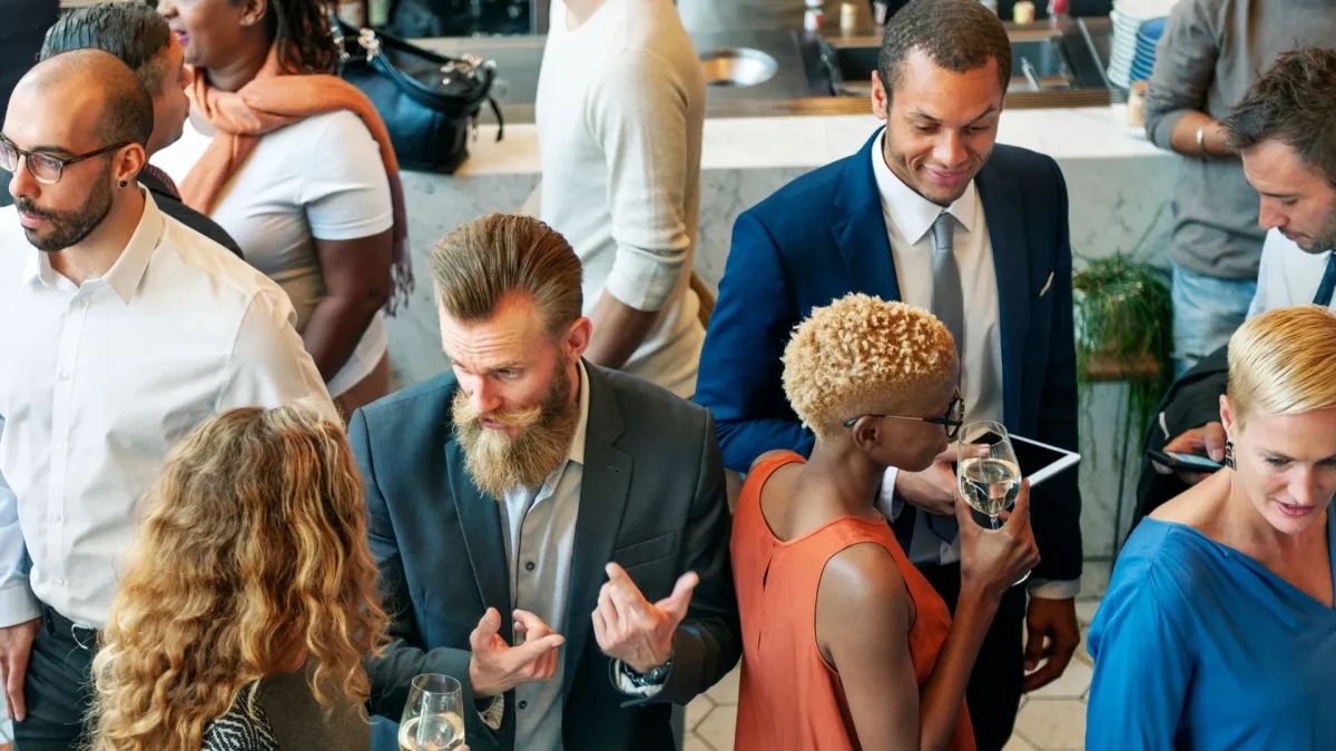 Fotografia em plano plongée (de cima para baixo) mostrando um grupo de pessoas conversando em um evento corporativo ou de networking. No centro, um homem de terno cinza escuro, cabelo penteado para trás e barba longa e clara fala gesticulando com as mãos para uma mulher de costas com cabelos cacheados. À direita, um homem negro de terno azul sorri enquanto olha para baixo. Próximo a ele, uma mulher de perfil, com cabelo loiro curto e crespo e blusa laranja, segura uma taça de vinho. O ambiente é bem iluminado, sugerindo um coquetel ou recepção social de negócios.