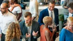 Fotografia em plano plongée (de cima para baixo) mostrando um grupo de pessoas conversando em um evento corporativo ou de networking. No centro, um homem de terno cinza escuro, cabelo penteado para trás e barba longa e clara fala gesticulando com as mãos para uma mulher de costas com cabelos cacheados. À direita, um homem negro de terno azul sorri enquanto olha para baixo. Próximo a ele, uma mulher de perfil, com cabelo loiro curto e crespo e blusa laranja, segura uma taça de vinho. O ambiente é bem iluminado, sugerindo um coquetel ou recepção social de negócios.