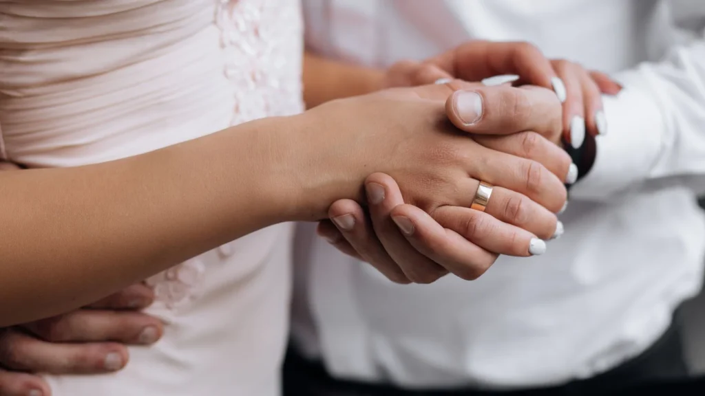 Fotografia em plano detalhe das mãos de um casal unidas e entrelaçadas, simbolizando o companheirismo e a renovação de votos. O homem, vestindo uma camisa branca, segura delicadamente a mão da mulher, destacando uma aliança dourada de casamento no dedo anelar dele. Ela usa um vestido claro com sutis detalhes em renda. Ao fundo, o outro braço do homem envolve suavemente a cintura da parceira em um abraço acolhedor.
