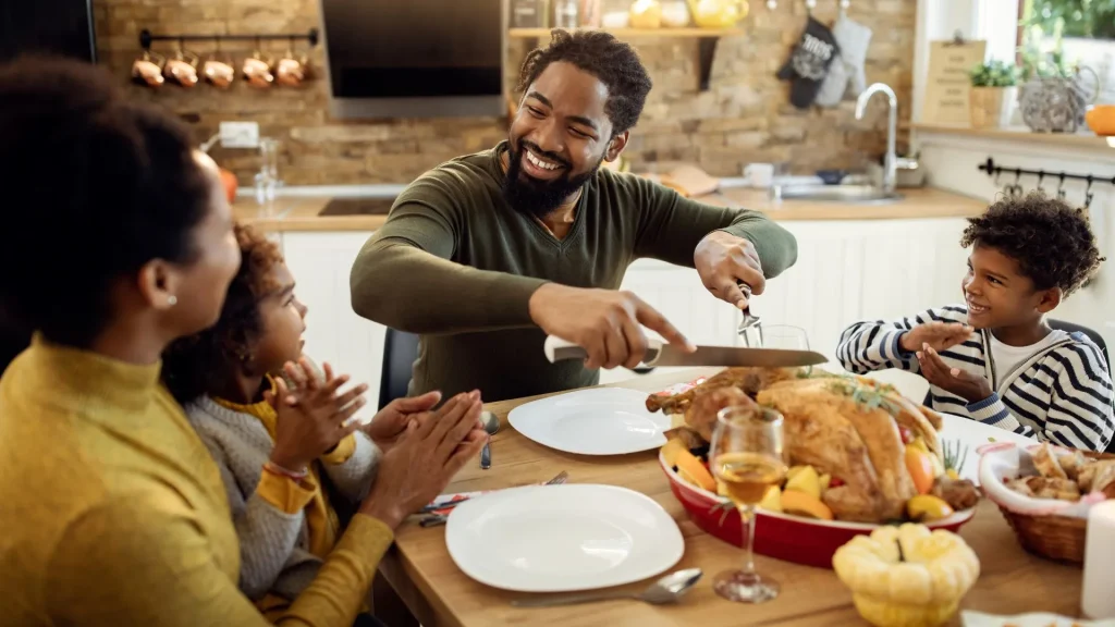 Uma família negra de quatro pessoas reunida ao redor de uma mesa de madeira em uma cozinha aconchegante, celebrando uma refeição festiva. Um homem sorridente e de barba, vestindo blusa verde de mangas compridas, está de pé cortando uma grande ave assada com uma faca e um garfo de servir. À direita, um menino de casaco listrado aplaude e sorri com entusiasmo. À esquerda, em primeiro plano, uma mulher de blusa amarela e uma menina também sorriem e aplaudem. Na mesa, há pratos brancos vazios, uma taça de vinho ou suco, uma cesta de pães e uma pequena abóbora decorativa. Ao fundo, vê-se uma bancada de cozinha moderna com tijolos expostos.