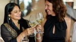 Um casal de mulheres sorri afetuosamente enquanto brinda com taças de vinho branco em um ambiente interno aconchegante. À esquerda, uma mulher de pele morena clara e longos cabelos pretos veste uma blusa preta com elegantes bordados dourados. À direita, sua parceira, uma mulher de pele clara com cabelos castanhos cacheados na altura dos ombros, usa um vestido preto de gola alta. Elas se olham com cumplicidade enquanto tocam as taças no centro da imagem, celebrando juntas.