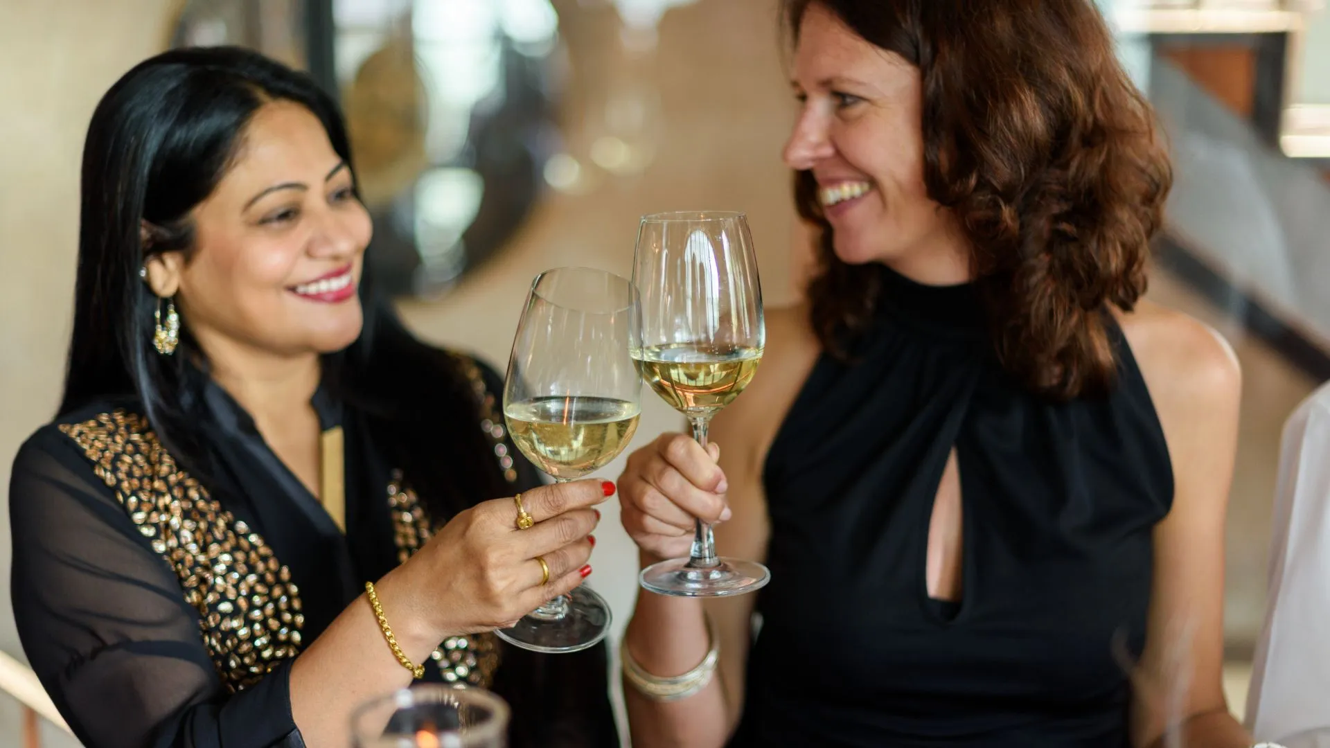 Um casal de mulheres sorri afetuosamente enquanto brinda com taças de vinho branco em um ambiente interno aconchegante. À esquerda, uma mulher de pele morena clara e longos cabelos pretos veste uma blusa preta com elegantes bordados dourados. À direita, sua parceira, uma mulher de pele clara com cabelos castanhos cacheados na altura dos ombros, usa um vestido preto de gola alta. Elas se olham com cumplicidade enquanto tocam as taças no centro da imagem, celebrando juntas.