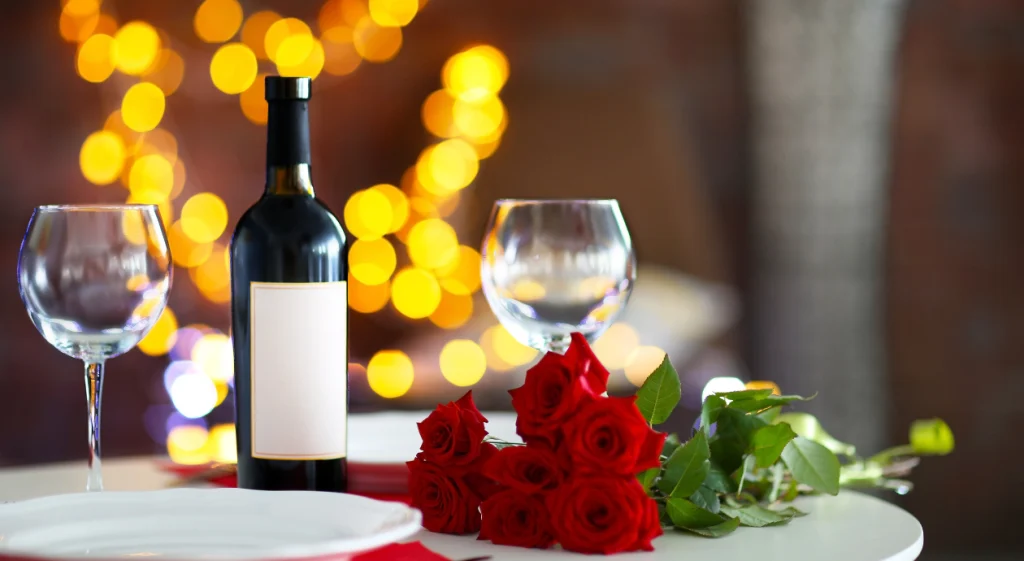 Mesa posta para jantar romântico com garrafa de vinho tinto, duas taças de cristal e buquê de rosas vermelhas, com luzes desfocadas ao fundo.