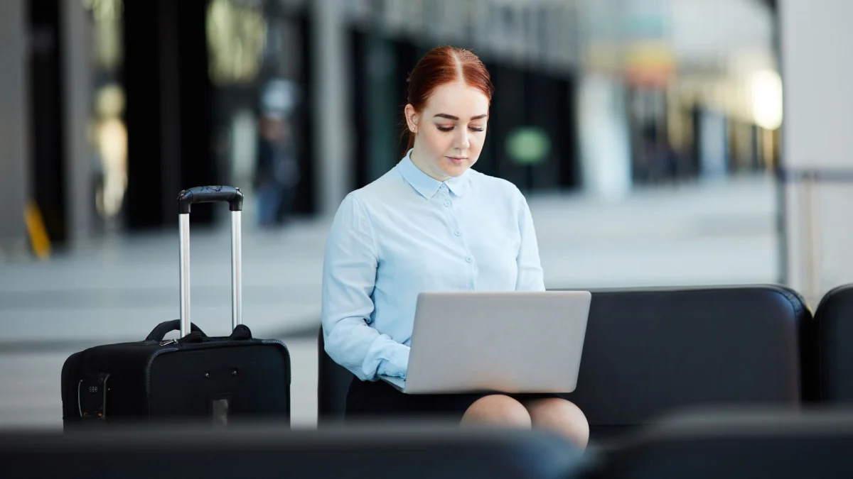 Uma mulher jovem de cabelos ruivos presos sentada em um banco de aeroporto ou saguão moderno, concentrada trabalhando em seu notebook portátil. Ao seu lado esquerdo, está uma mala de viagem de rodinhas preta. A imagem exemplifica o conceito de "bleisure" e "workation", onde o trabalho remoto se mistura com viagens de negócios e lazer.