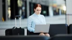 Uma mulher jovem de cabelos ruivos presos sentada em um banco de aeroporto ou saguão moderno, concentrada trabalhando em seu notebook portátil. Ao seu lado esquerdo, está uma mala de viagem de rodinhas preta. A imagem exemplifica o conceito de "bleisure" e "workation", onde o trabalho remoto se mistura com viagens de negócios e lazer.
