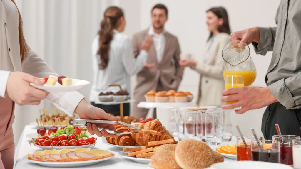 Uma pessoa se serve de lanches em uma mesa de buffet de coffee break corporativo, enquanto colegas de terno conversam ao fundo. Há croissants, suco de laranja, frutas e muffins.