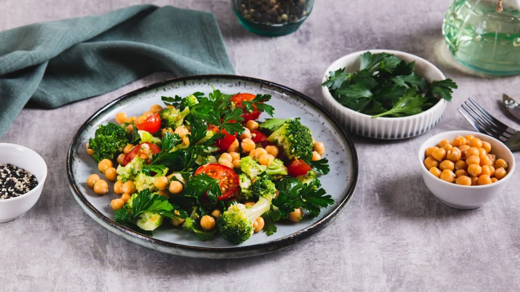 Vista superior de um prato com salada de grão-de-bico, buquês de brócolis cozido, tomates-cereja cortados ao meio e folhas de salsa fresca. Ao redor do prato, sobre uma mesa cinza, há pequenas tigelas com salsa, grão-de-bico extra e sementes de gergelim.