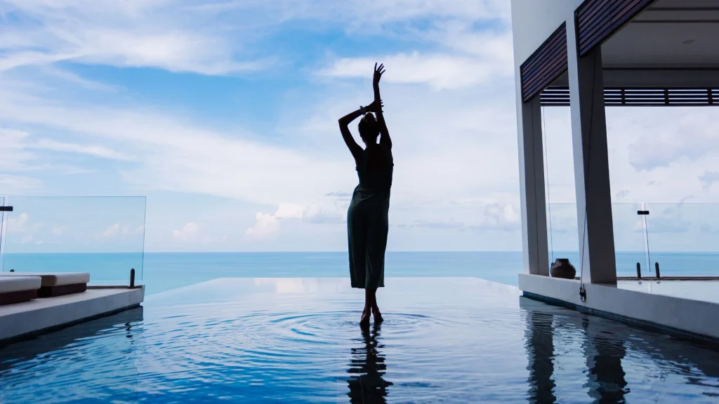 Silhueta elegante de uma mulher posando graciosamente em uma piscina de borda infinita em um resort de luxo, com vista para o oceano sob um céu azul.