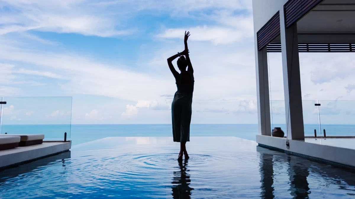 Silhueta elegante de uma mulher posando graciosamente em uma piscina de borda infinita em um resort de luxo, com vista para o oceano sob um céu azul.