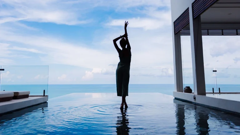 Silhueta elegante de uma mulher posando graciosamente em uma piscina de borda infinita em um resort de luxo, com vista para o oceano sob um céu azul.