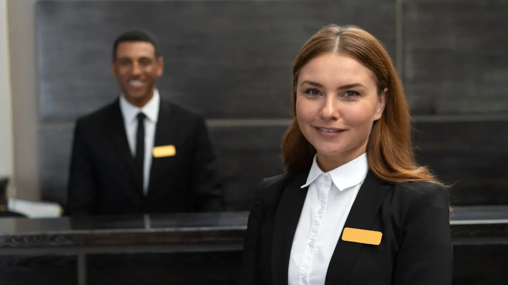 Dois funcionários uniformizados na recepção de um hotel: um homem sorrindo atrás do balcão e uma mulher sorrindo na frente do balcão. Ambos estão representando o serviço de concierge de viagens de luxo.