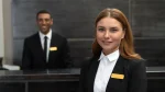 Dois funcionários uniformizados na recepção de um hotel: um homem sorrindo atrás do balcão e uma mulher sorrindo na frente do balcão. Ambos estão representando o serviço de concierge de viagens de luxo.