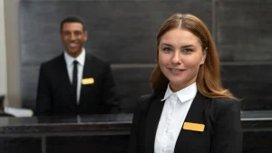 Dois funcionários uniformizados na recepção de um hotel: um homem sorrindo atrás do balcão e uma mulher sorrindo na frente do balcão. Ambos estão representando o serviço de concierge de viagens de luxo.