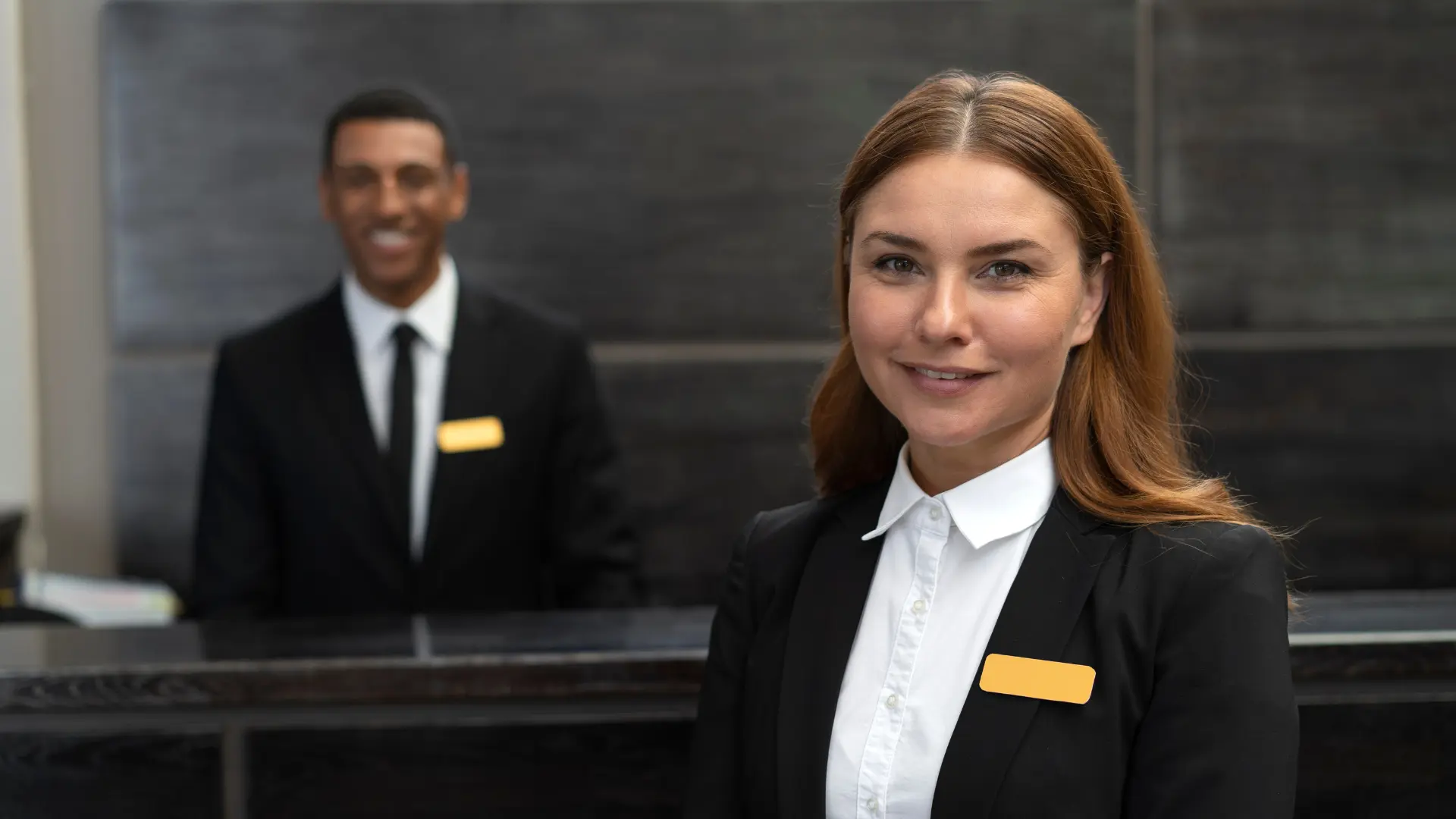 Dois funcionários uniformizados na recepção de um hotel: um homem sorrindo atrás do balcão e uma mulher sorrindo na frente do balcão. Ambos estão representando o serviço de concierge de viagens de luxo.
