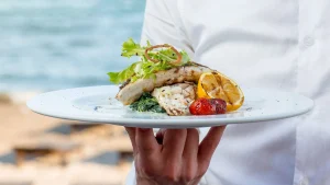Garçom segura um prato com peixe grelhado, tomate assado e limão siciliano à beira-mar. A refeição é decorada com folhas verdes frescas.
