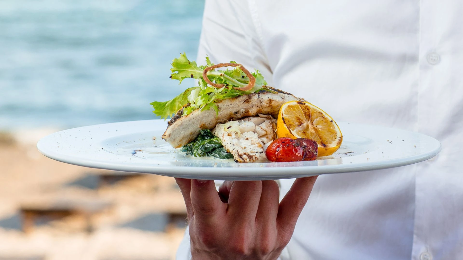 Garçom segura um prato com peixe grelhado, tomate assado e limão siciliano à beira-mar. A refeição é decorada com folhas verdes frescas.