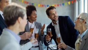 Colegas de trabalho sorriem e conversam segurando taças de espumante em uma celebração de escritório. Bandeirinhas coloridas decoram o ambiente ao fundo.
