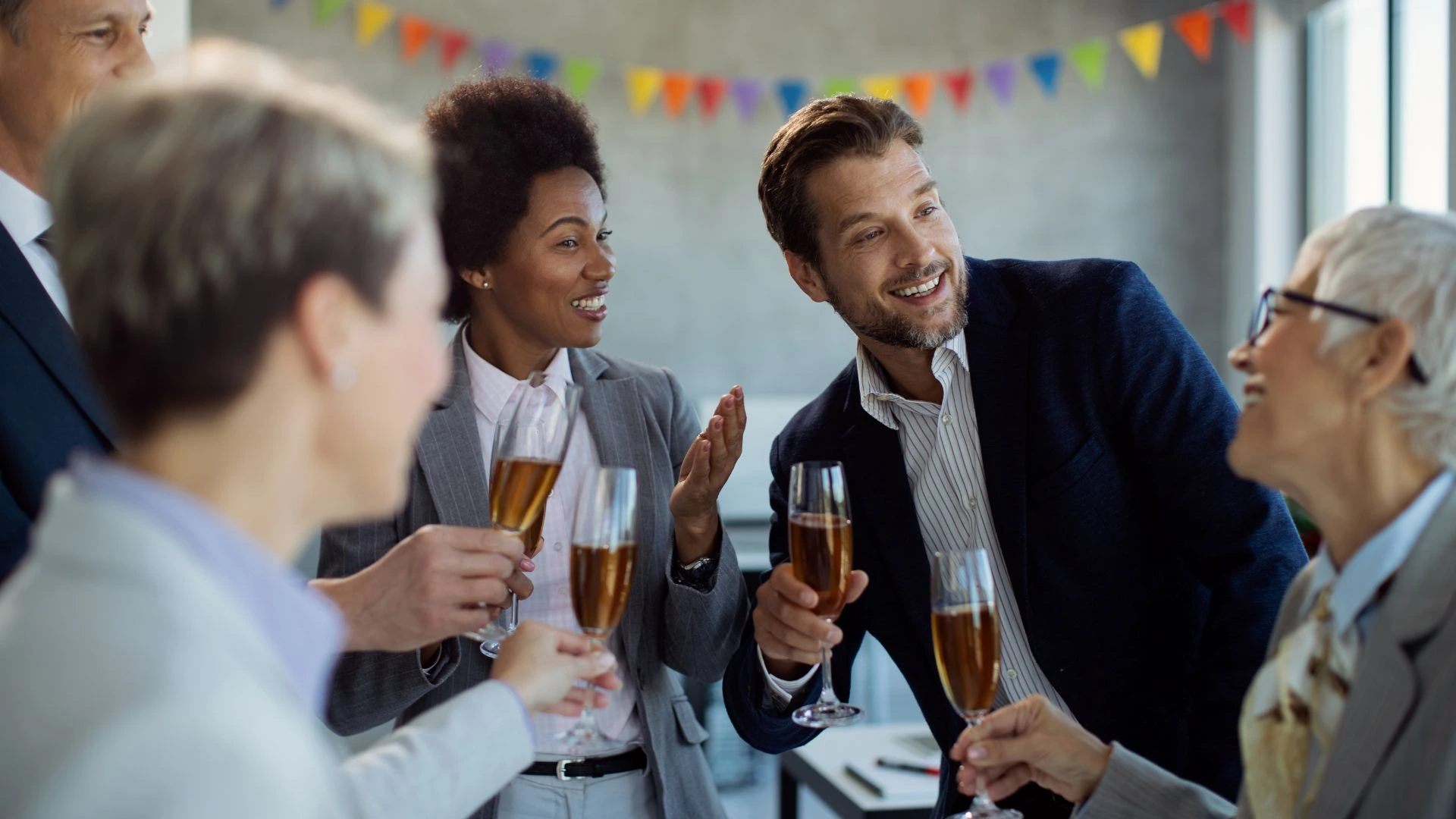 Colegas de trabalho sorriem e conversam segurando taças de espumante em uma celebração de escritório. Bandeirinhas coloridas decoram o ambiente ao fundo.