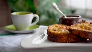 Fatias de pão douradas em um prato branco, ao lado de uma xícara de café. Um potinho com colher aparece desfocado ao fundo.
