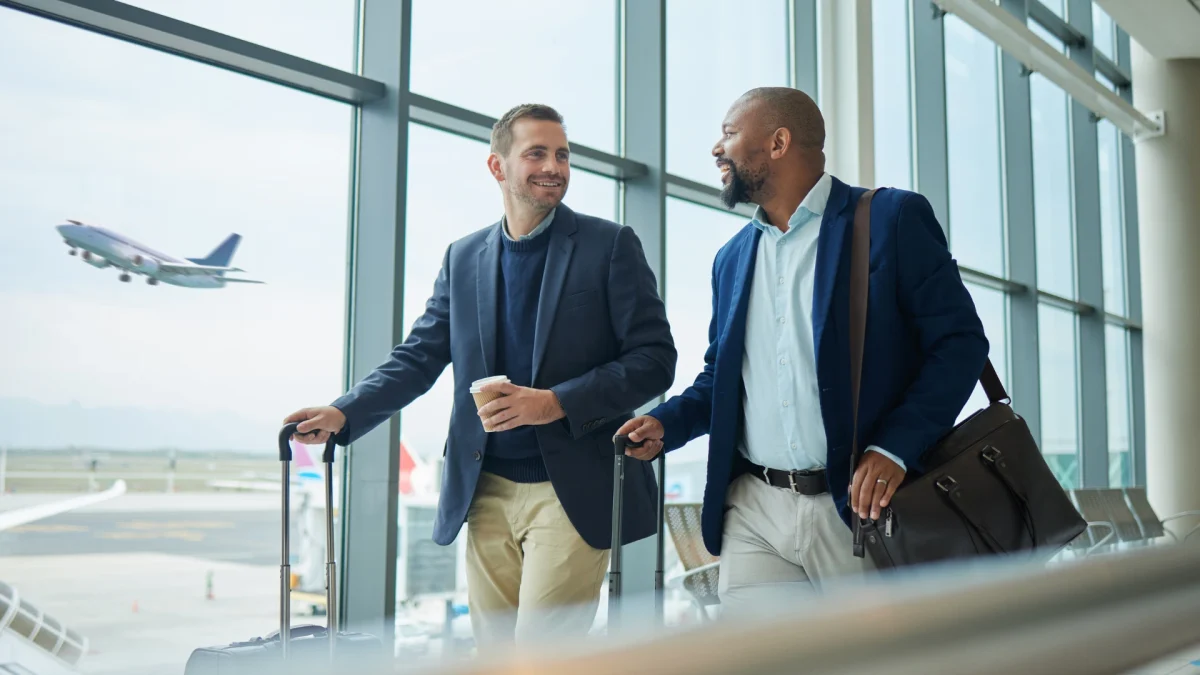 Dois executivos sorridentes conversam em um saguão de aeroporto, puxando malas de viagem. Ao fundo, um avião decola sob janelas amplas e iluminadas.