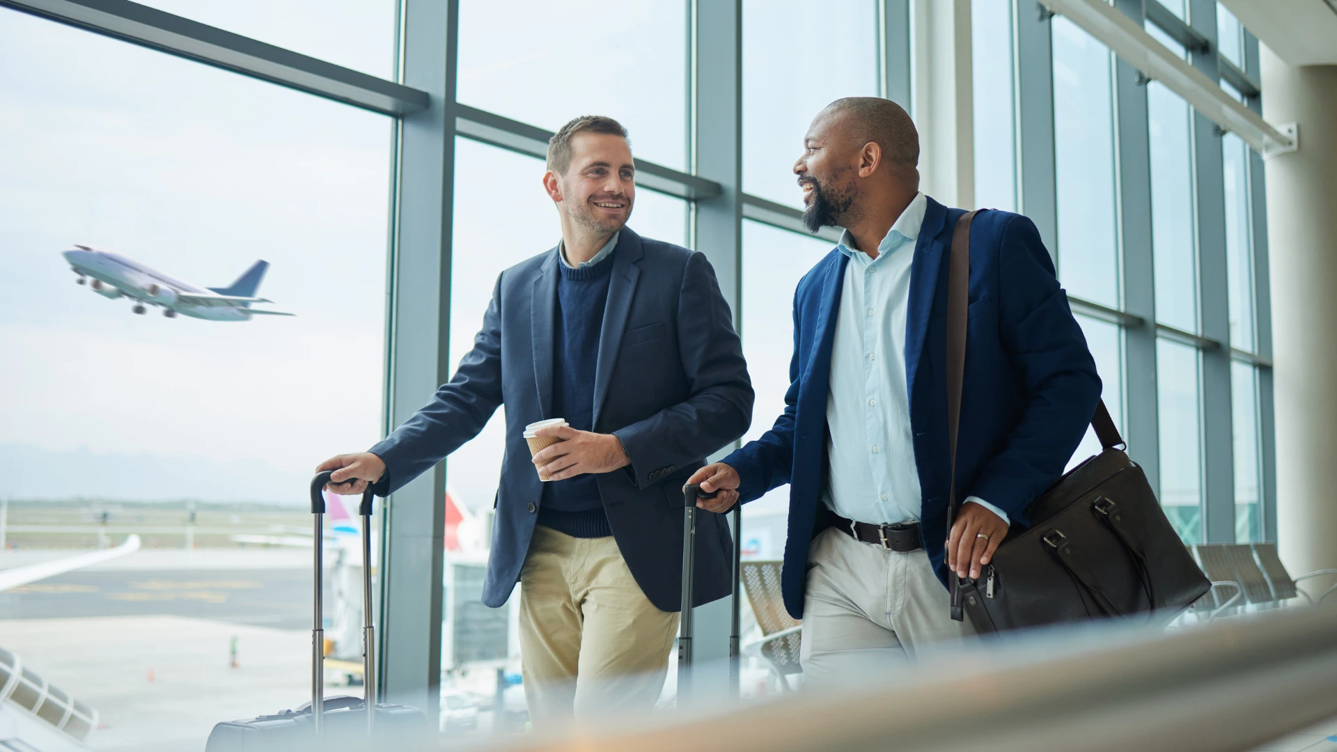 Dois executivos sorridentes conversam em um saguão de aeroporto, puxando malas de viagem. Ao fundo, um avião decola sob janelas amplas e iluminadas.