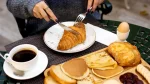 Mãos cortam um croissant em uma farta mesa verde de café da manhã ao ar livre. O banquete inclui café preto, ovo cozido, panquecas e pães.