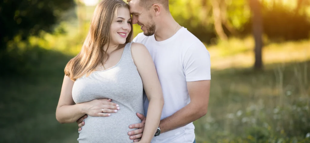 Homem abraça por trás a esposa grávida no parque, enquanto ambos apoiam as mãos carinhosamente sobre a sua barriga.