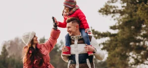 Família com roupas de inverno no parque: pai carrega a filha nos ombros enquanto a mãe toca na mão da criança sorrindo.