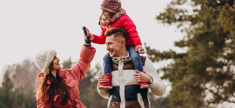 Família com roupas de inverno no parque: pai carrega a filha nos ombros enquanto a mãe toca na mão da criança sorrindo.