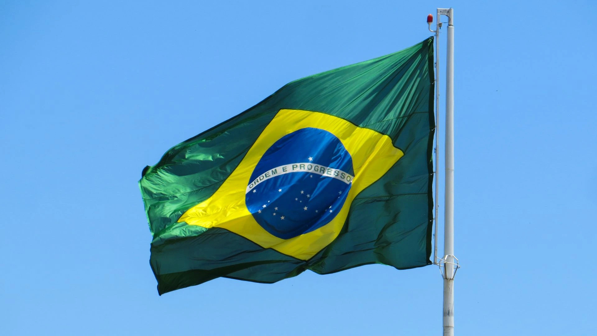 Bandeira do Brasil tremula ao vento em um mastro, sob céu azul e claro. O símbolo verde, amarelo e azul representa o país.