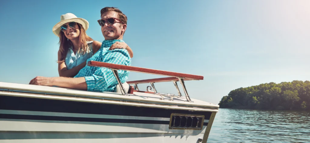 Casal sorridente usando óculos de sol passeia de barco em um dia ensolarado, com vegetação e água ao fundo.