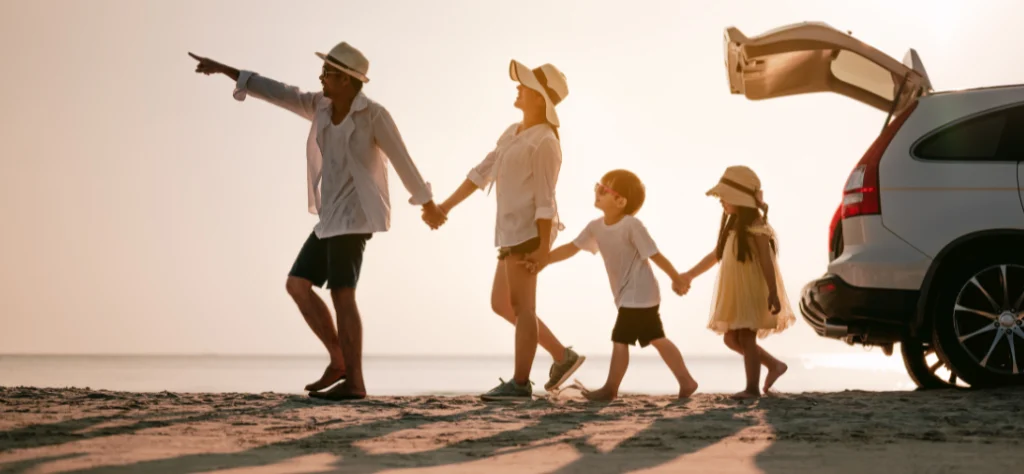 Família de quatro pessoas caminha de mãos dadas na praia ao pôr do sol, ao lado de um carro com o porta-malas aberto.