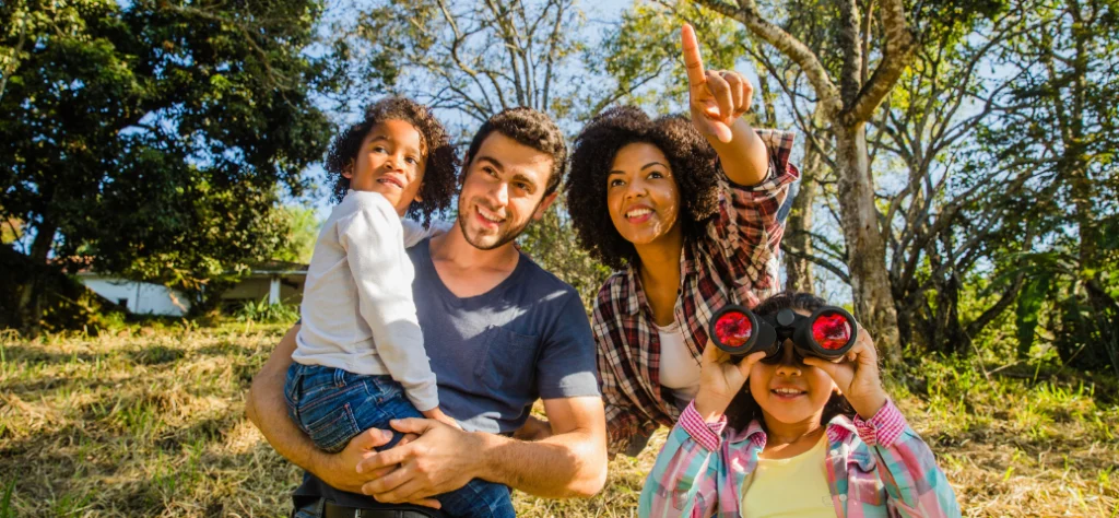 Família em passeio ao ar livre: mulher aponta para o alto e menina sorri usando binóculos em meio à natureza.