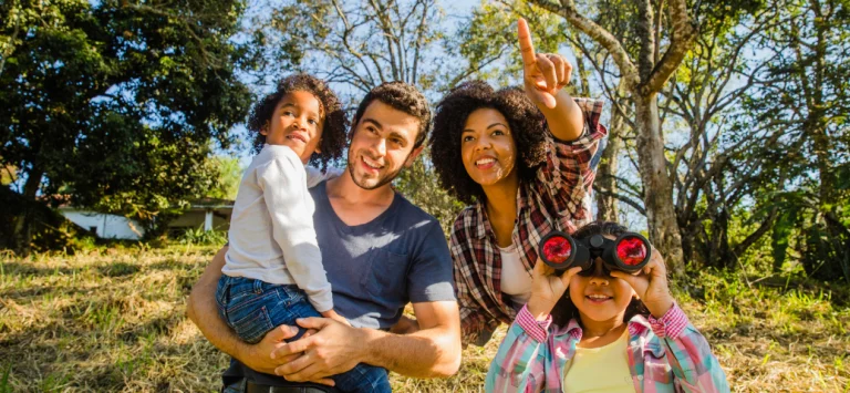 Família em passeio ao ar livre: mulher aponta para o alto e menina sorri usando binóculos em meio à natureza.
